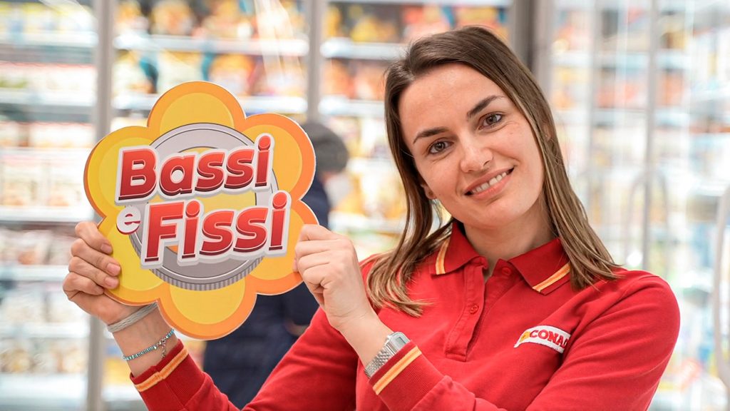 Dipendente Conad sorride mostrando cartello "Bassi e Fissi" in supermercato
