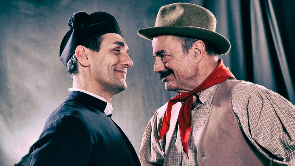 Attori in costume da Don Camillo e Peppone si guardano sorridendo sul set