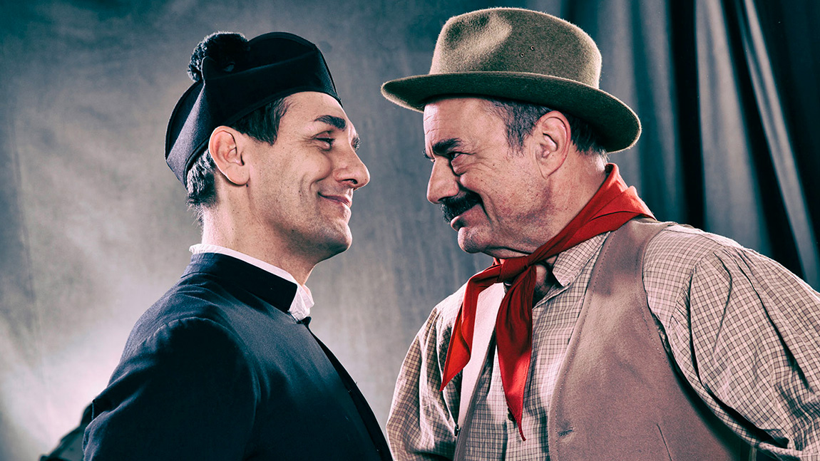 Attori in costume da Don Camillo e Peppone si guardano sorridendo sul set