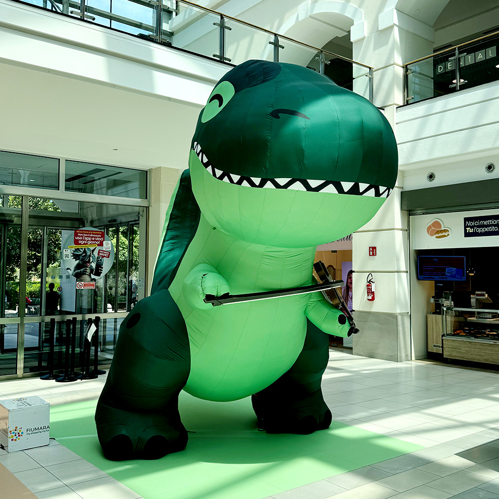 Un grande dinosauro gonfiabile verde si trova nel centro commerciale Fiumara, con in mano un violino e un archetto.